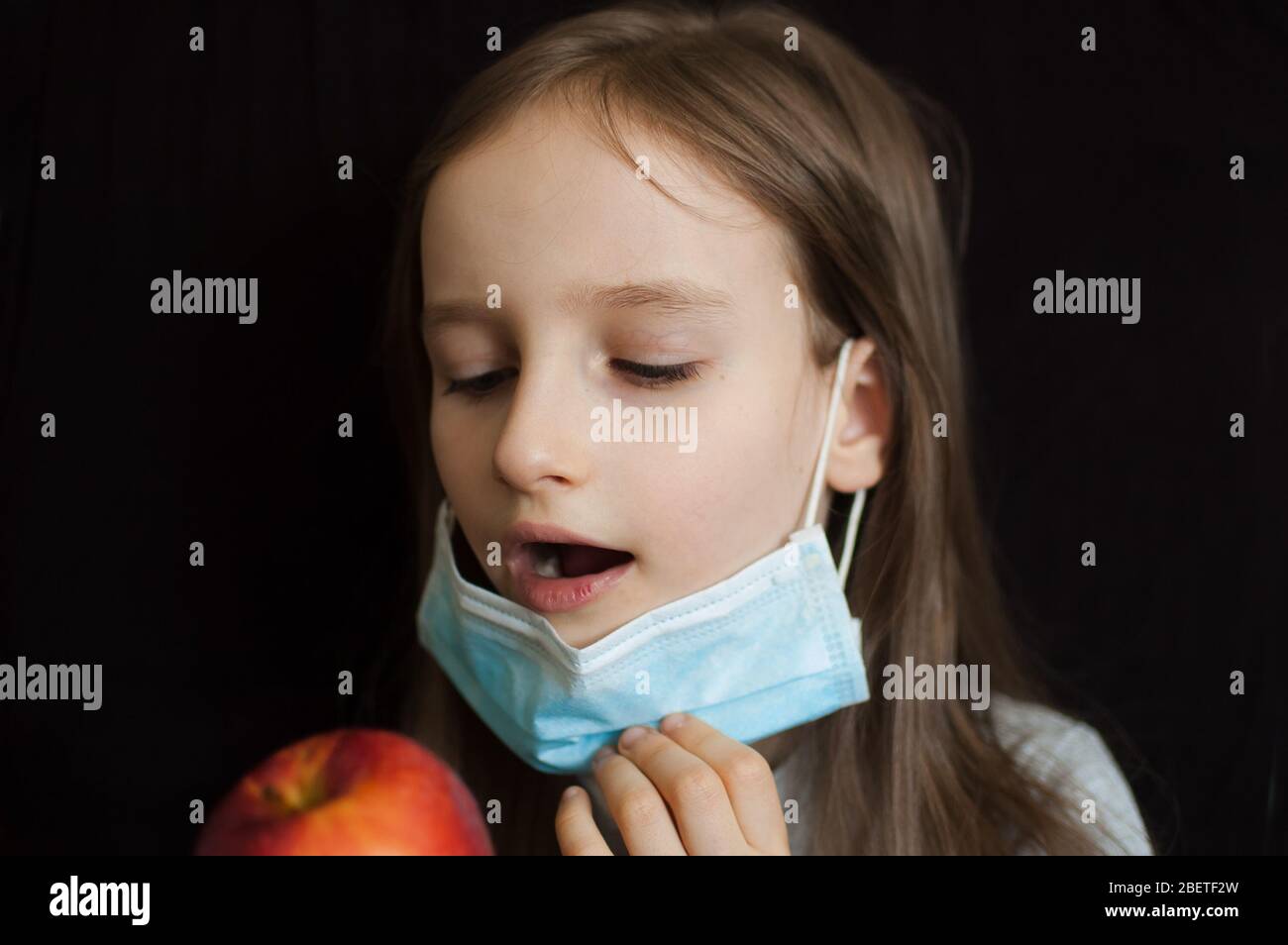 Kleines blondes Mädchen mit blauen Augen, das eine Einwegmaske zum Schutz von Coronavirus Covid-19 trägt, isst einen roten apfel, um ihr Immunsystem anzukurbeln Stockfoto