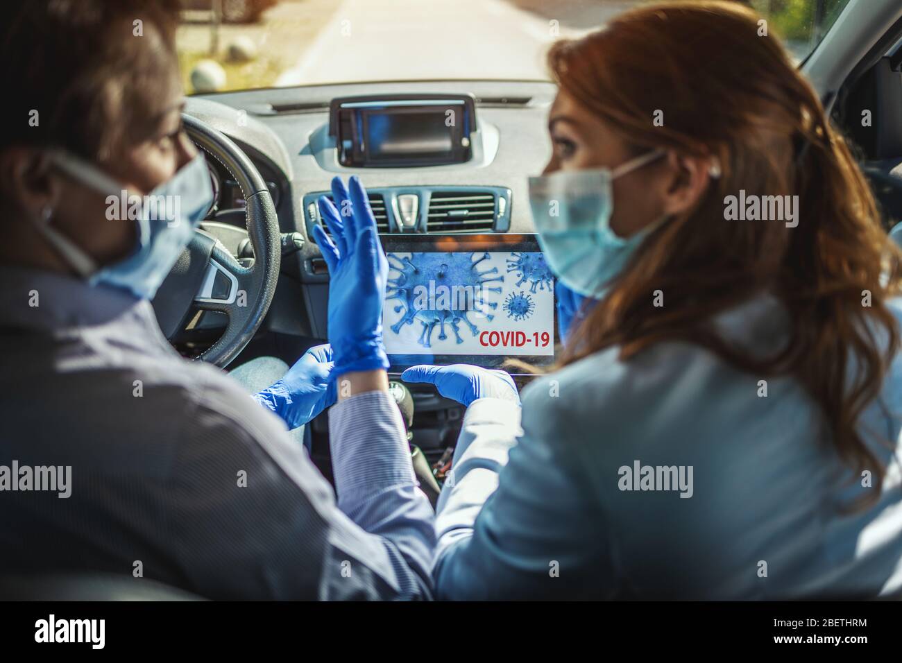 Junge Geschäftsfrauen mit Schutzmasken im Gesicht sitzen in einem Auto und reden über globale Krise. Sie zeigen sich gegenseitig eine Animati Stockfoto