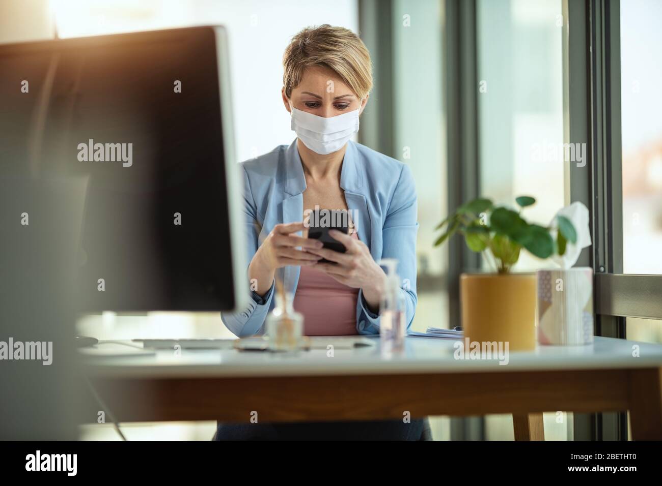Geschäftsfrau in einer medizinischen Schutzmaske arbeitet von zu Hause aus am Computer, mit dem Smartphone, während der Selbstisolierung und Quarantäne, um Infektionen zu vermeiden Stockfoto