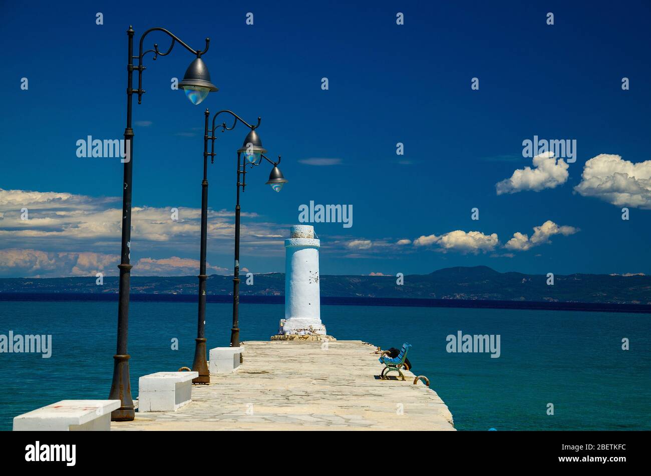 Weißer Pier Steg mit Bank, Lampenpfosten und Leuchtturm auf blauem Paradieswasser des Golfs von Toroneos kolpos, blauer Himmel, weiße Wolken über der Halbinsel Sithonia i Stockfoto