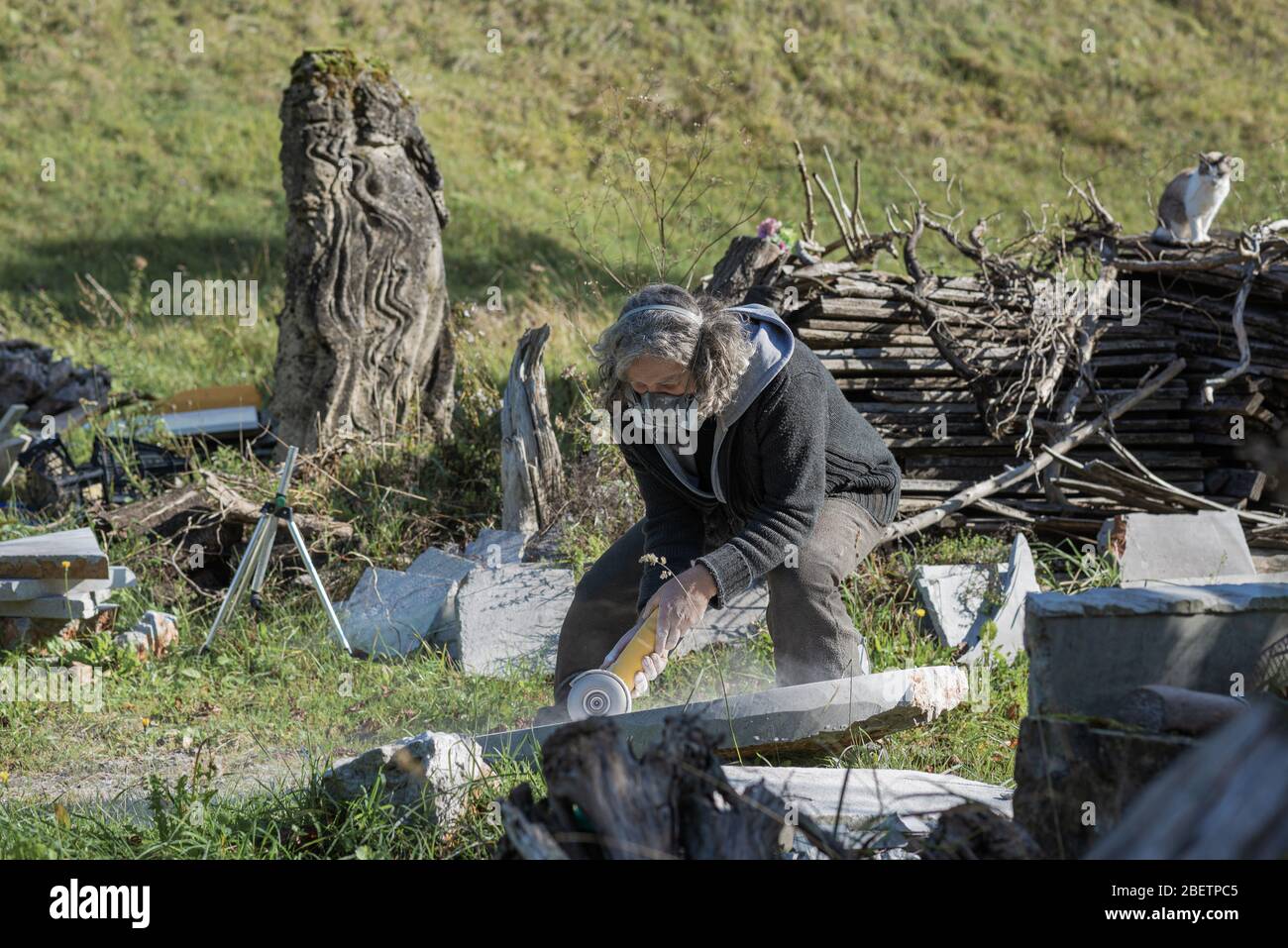 Senior Künstler trägt Schutzmaske Arbeit an seinem neuesten Projekt, Polieren Stein mit einem Schleifer draußen mit seinen Werken und materia umgeben Stockfoto