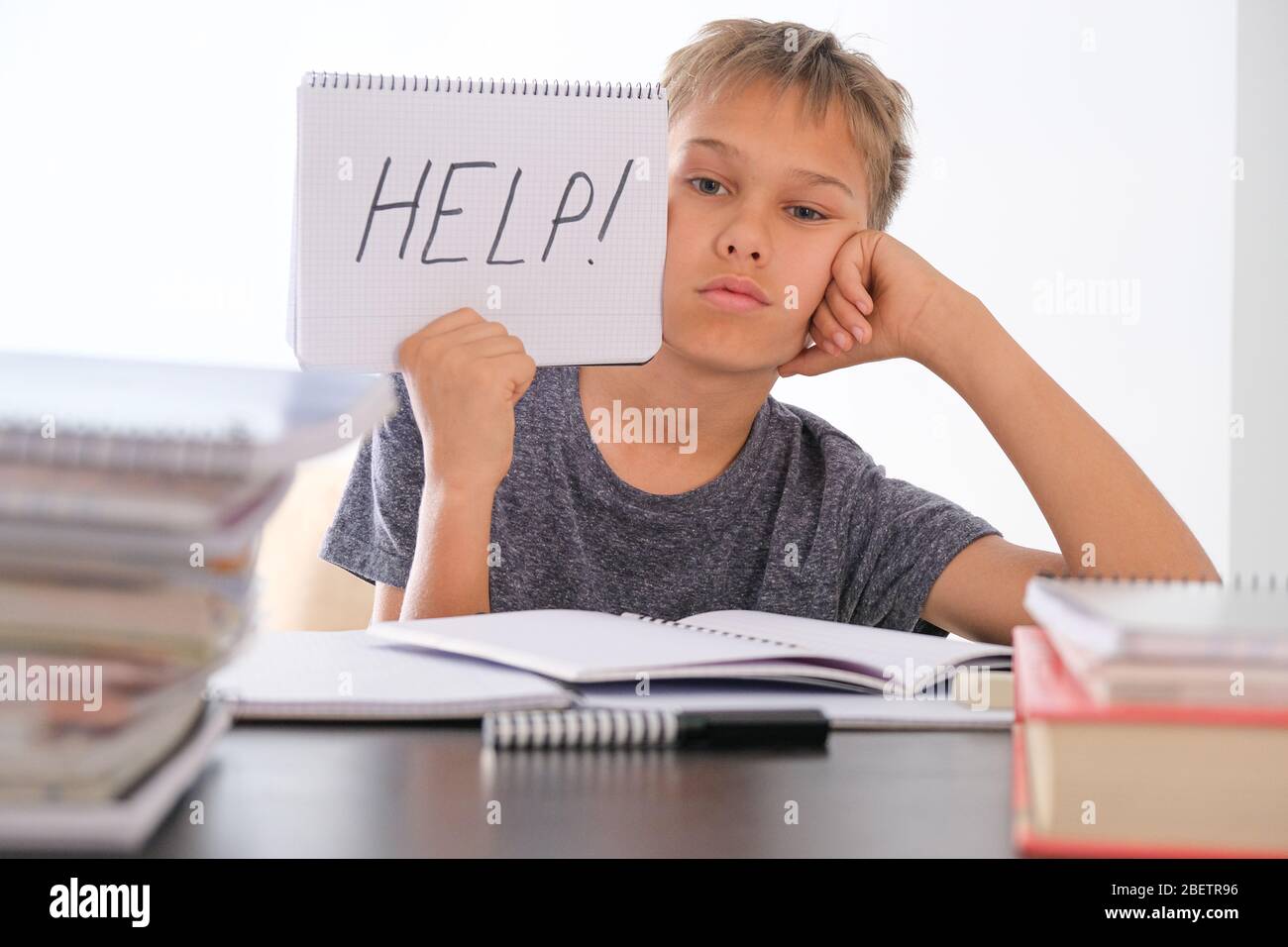 Verärgert müde preteen Kind sitzt am Tisch, seine Hausaufgaben unter Haufen von Büchern. Word Help ist auf offenem Notizbuch geschrieben Stockfoto