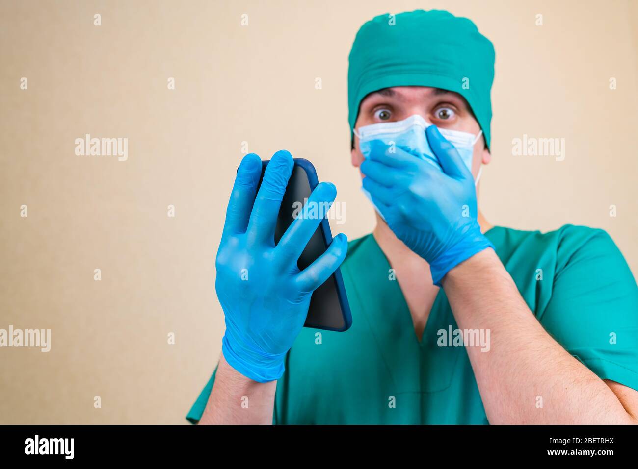 Doktor in schützender medizinischer Maske schockiert aussehende Nachrichten in seinem Handy in der Hand isoliert auf glattem Hintergrund mit Kopierraum für Text Stockfoto