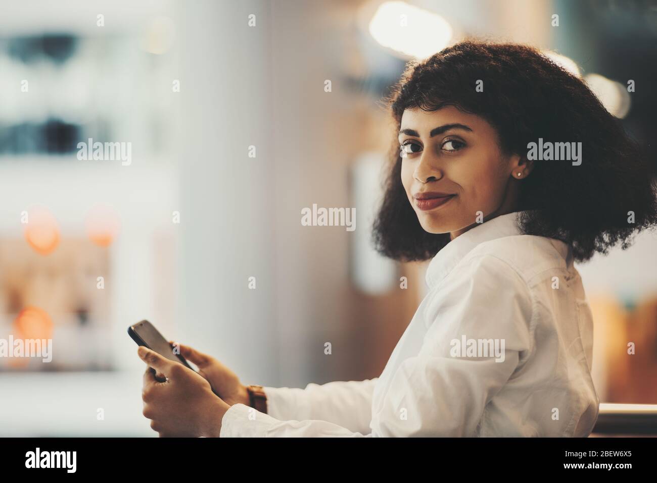 Ein Innenporträt einer jungen blendenden afroamerikanischen Frau in einem weißen Hemd mit Handy und lockigen Afro-Haaren, die die Kamera anschaut; eine charmante Bira Stockfoto