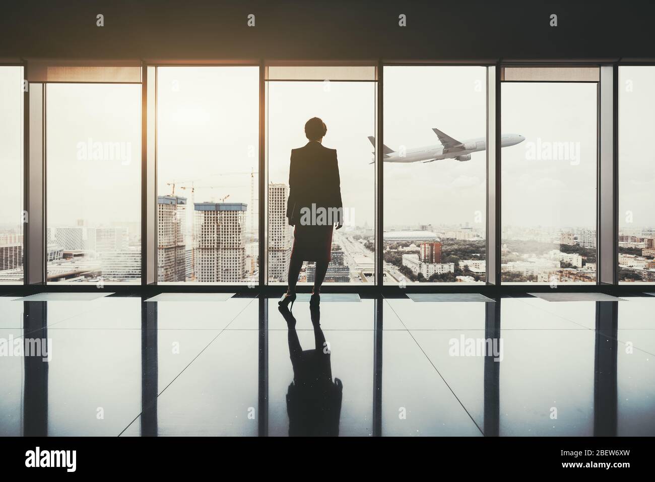 Eine Silhouette einer erfolgreichen Unternehmerin, die drinnen von einem luxuriösen Interieur mit einem reflektierenden Marmorboden steht, und einem Flugzeug, das an Höhe gewinnt Stockfoto