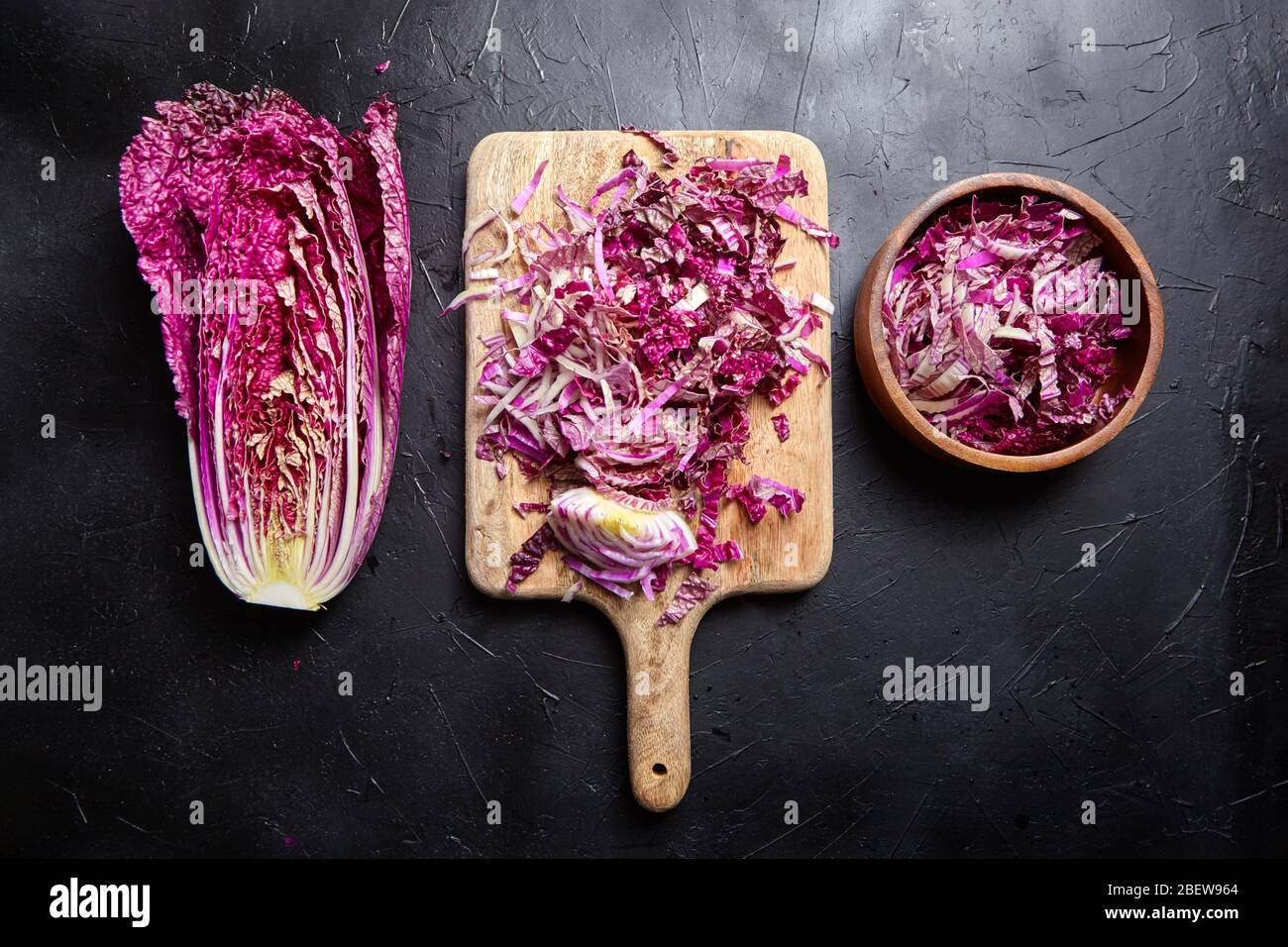 Gehackter Rotchinesischer Kohl (Lila Wombok) auf Holzbrett und Schüssel auf Steintisch. Halber Rotkohl auf schwarzem Hintergrund, Draufsicht. Kochen Stockfoto