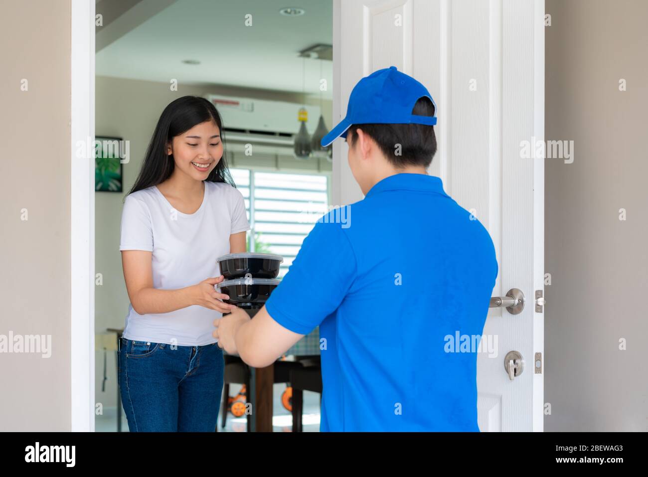 Asiatische Lieferung junger Mann in blauem, gleichmäßigem Lächeln und mit Essenskisten im Vorderhaus und Asian Woman, die eine Lieferung von Lebensmittelkisten aus Deliverym annehmen Stockfoto