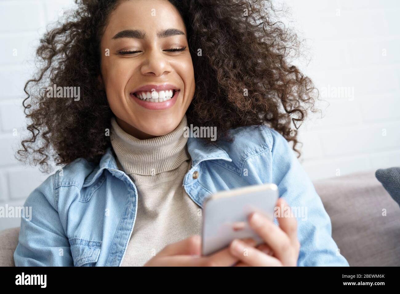 Fröhliche Afro amerikanische junge Frau lacht mit Handy-Apps zu Hause. Stockfoto