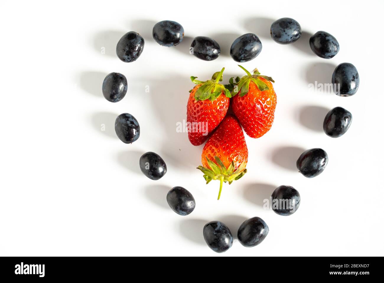 Schwarze Trauben in Form eines Liebesherzens mit drei Erdbeeren in der Mitte vor einem schlichten weißen Hintergrund Stockfoto