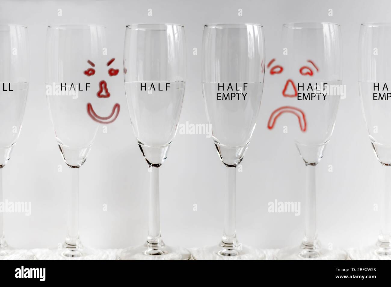 Sechs Weingläser, halb voll mit gezeichneten traurigen und glücklichen Gesichtern im Hintergrund, die Konzept von Glas halb voll oder Glas halb leer darstellen. Stockfoto