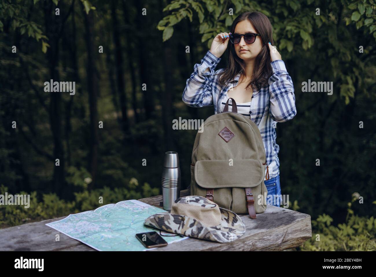 Mädchen in Sonnenbrille im Wald mit Rucksack. Touristenkarte, Thermoskanne auf einem Holztisch. Stockfoto