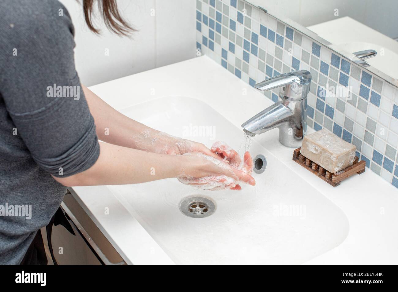 Frau, die ihre Hand mit grüner Seife vor ihrem weißen Waschbecken wäscht. Wasser fließt durch den silbernen Wasserhahn. An der Wand sind kleine Fliesen Stockfoto