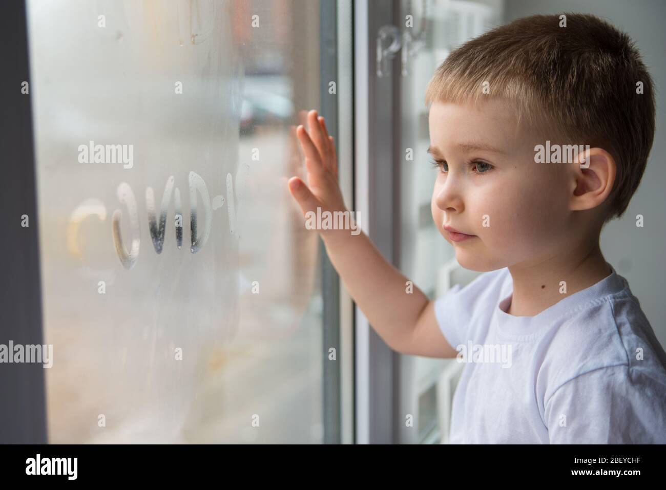 Cute 4 Jahre Old Boy Blick durch das Fenster und halten Sie seine Hand auf das Glas mit Wort Covid 19 Stockfoto