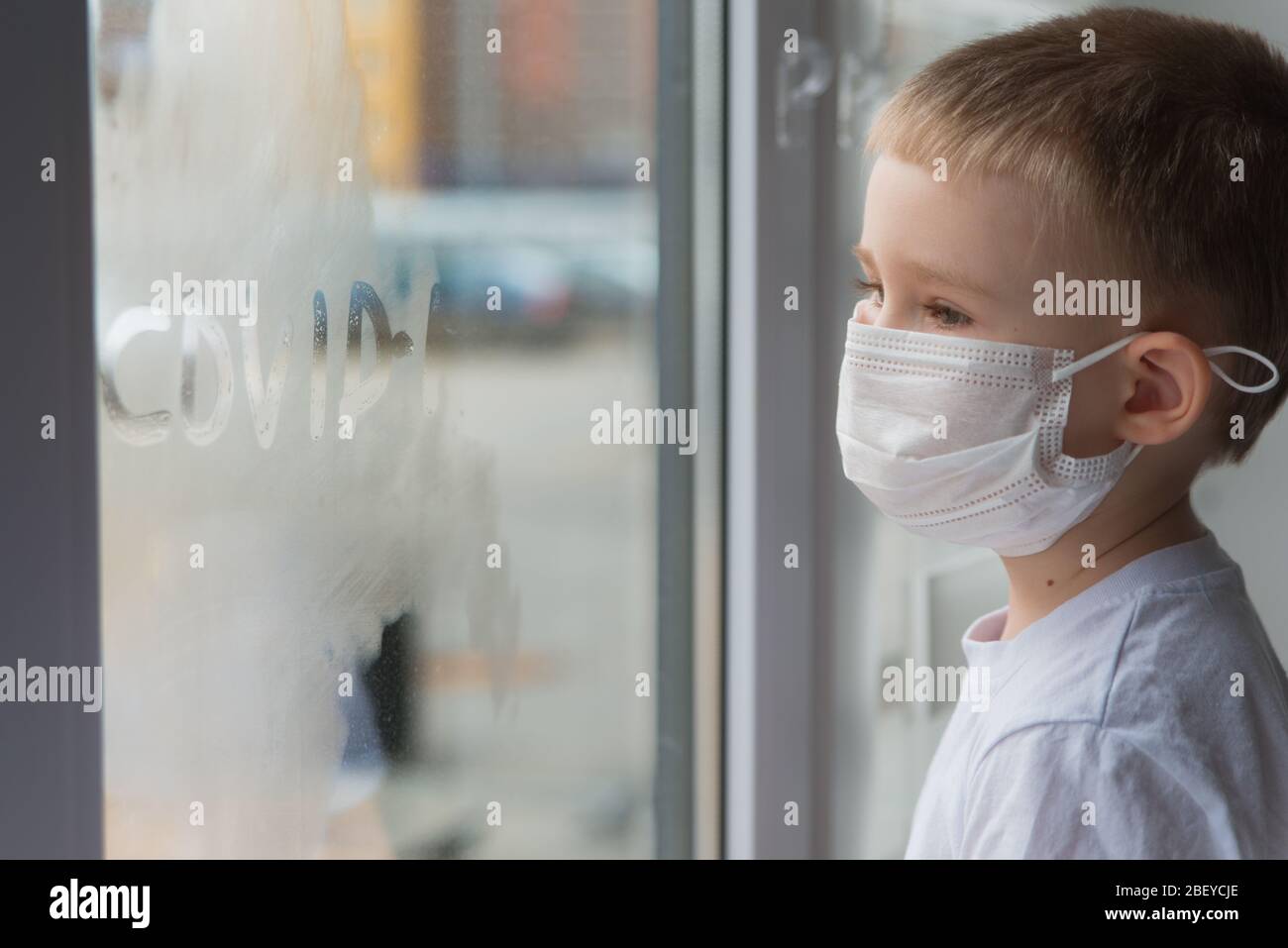 Niedliche 4 Jahre alt Junge in medizinischen Maske Blick durch das Fenster mit Wort Covid 19 Stockfoto