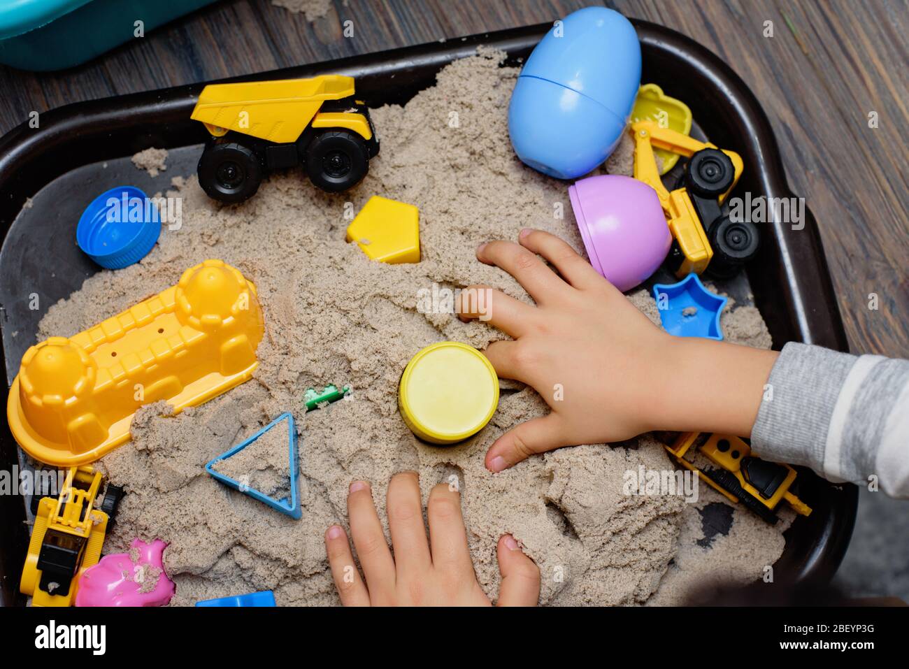 Kind spielt mit kinetischem Sand und Spielzeugbaumaschinen. Hand des Kindes in Sand Nahaufnahme. Flach liegend, Draufsicht. Indoor-Tischspiel. Kreativitätsspiel Stockfoto