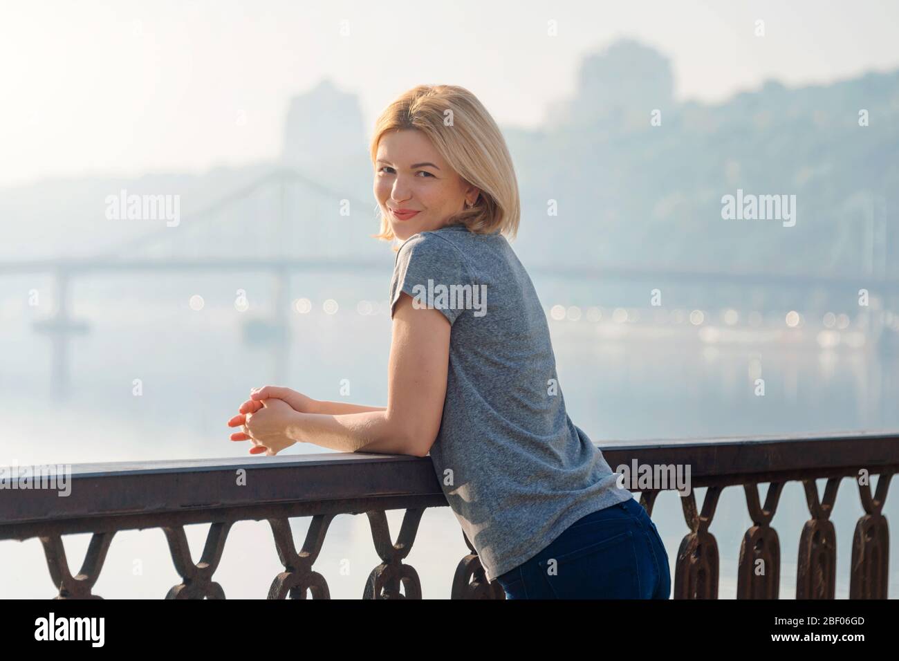 Junge blonde Frau genießt Blick auf die Stadt von der Brücke. Schöne Frau, die Zeit auf dem Flussufer bei Sonnenuntergang verbringt. Weiche Hintergrundbeleuchtung. Lifestyle Relax Stockfoto