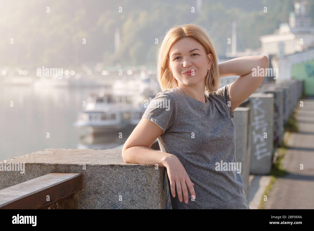Junge blonde Frau genießt Blick auf die Stadt von der Brücke. Schöne Frau, die Zeit auf dem Flussufer bei Sonnenuntergang verbringt. Weiche Hintergrundbeleuchtung. Lifestyle Relax Stockfoto