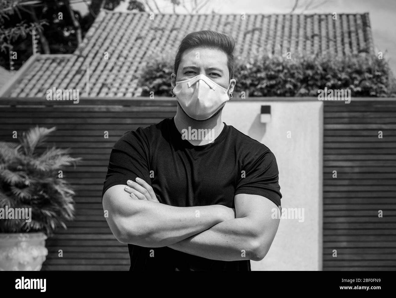 Porträt eines erwachsenen Mannes mit verschränkten Armen, der während der Quarantäne zu Hause eine weiße Gesichtsmaske trägt. Schwarzweiß-Foto. Stockfoto