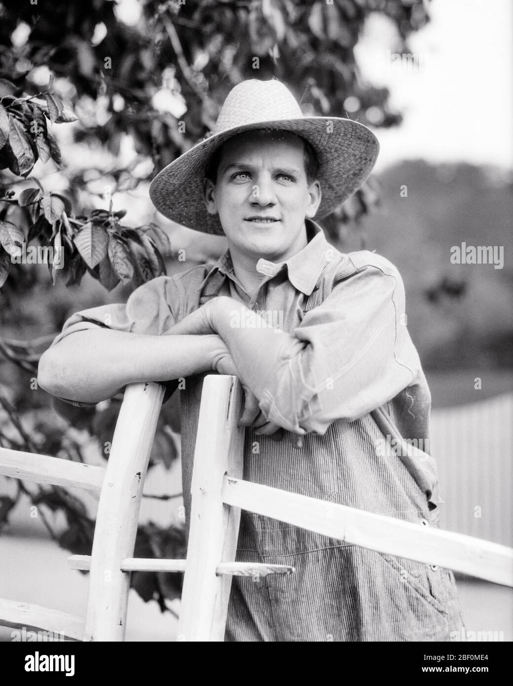 1920ER JAHRE PORTRÄT DES NACHDENKLICHEN LANDWIRT TRÄGT TRADITIONELLE STROHHUT UND BIB OVERALLS SCHIEFEN ARME AUF HOLZTOR GEKREUZT - F3334 HAR001 HARS LANDWIRTSCHAFT B & W RUHE TOR ZIELE FÄHIGKEIT BERUF FÄHIGKEITEN UND KARRIERE BAUERN FÜHRUNG BERUFE VERBINDUNG PAUSE BIB WACHSTUM MITTE-ERWACHSENE MITTE-ERWACHSENE MANN SCHWARZ UND WEISS KAUKASISCHEN ETHNIZITÄT HAR001 ALTMODISCH Stockfoto