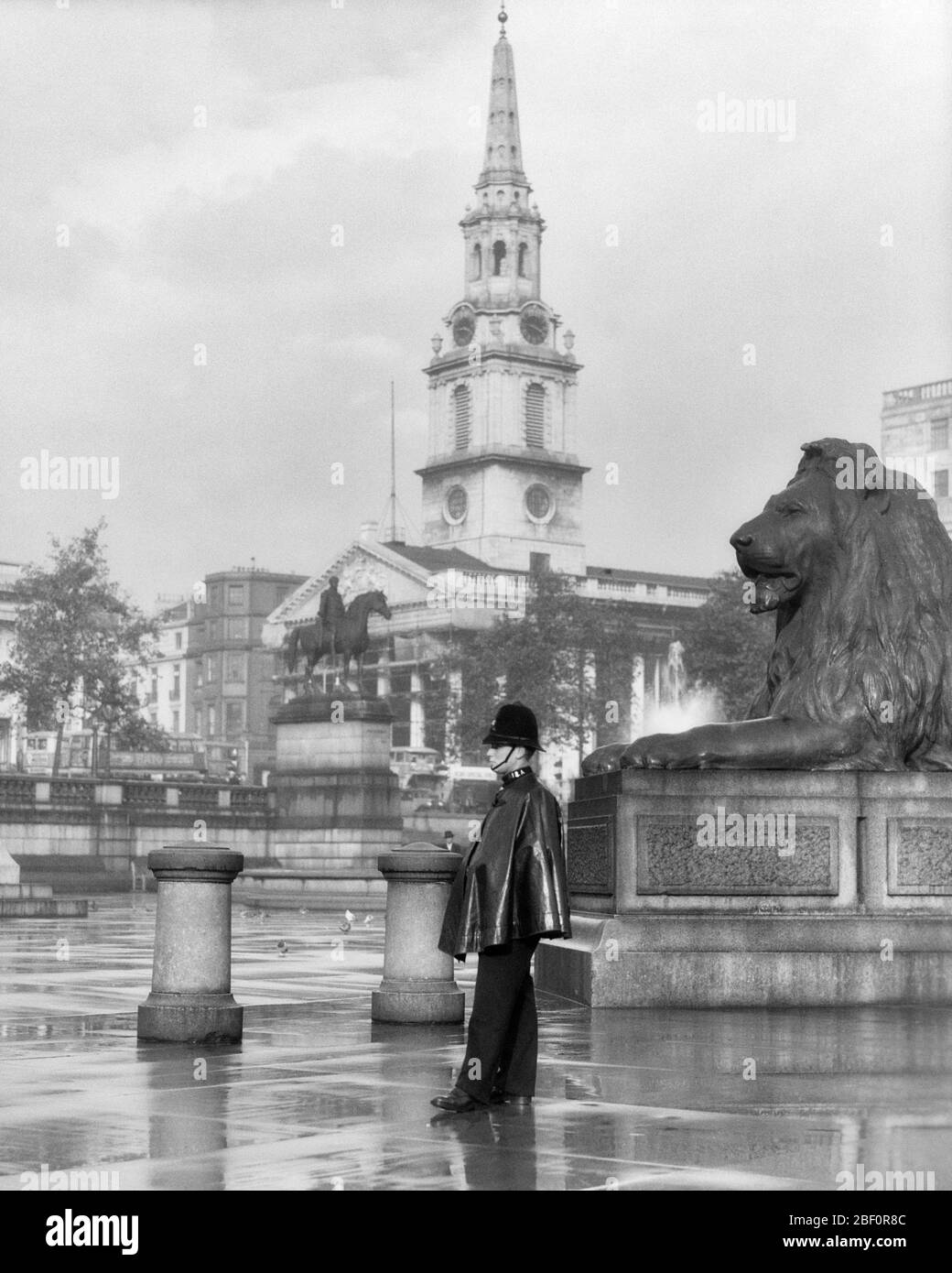 1930ER REGENTAG LONDON BOBBY STEHT IN DER NÄHE DER LION STATUE TRAFALGAR SQUARE ST. MARTIN-IN-THE-FIELDS ANGLIKANISCHE KIRCHE IN B/G ENGLAND - R7643 HAR001 HARS RAINY LION PERSONS TRADITIONELLE MÄNNER RAINING OFFICER NEXT ENGLISCH B&W BRITAIN COP SCHÜTZEN UND DIENEN HOCHWINKEL SCHUTZ STÄRKE MUT STOLZ AUTORITÄT BERUFE UNIFORMEN KONZEPTIONELLE BOBBY ST. OFFIZIERE POLIZISTEN ANGLIKANISCHE STADT WESTMINSTER COPS KOPFBEDECKUNG MITTE ERWACHSENE MITTE ERWACHSENE MANN JUNGE ERWACHSENE MANN ABZEICHEN ABZEICHEN SCHWARZ UND WEISS KAUKASISCHEN ETHNIZITÄT GROSSBRITANNIEN HAR001 IKONISCHE ALTMODISCHE TOURISMUS TRAFALGAR VEREINIGTES KÖNIGREICH Stockfoto