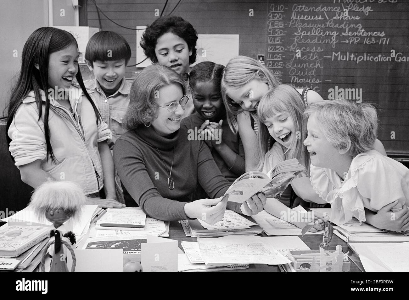 1970S 1980S VIELFÄLTIGE GRUPPE VON SCHULKINDERN JUNGEN UND MÄDCHEN UM LEHRER HALTEN OFFENES BUCH AN IHREM SCHREIBTISCH - S21516 HAR001 HARS HER ASIAN OLD TIME FUTURE NOSTALGIE LESEN TAFEL ALTE MODE JUGENDLICH DIVERSE LEHRER KOMMUNIKATION LACHEN SPANISCH INFORMATIONEN ERFREUT FREUDE LIFESTYLE FRAUEN KOPIEREN RAUM FREUNDSCHAFT HALBE LÄNGE DAMEN PERSONEN INSPIRATION MÄNNER VERTRAUEN B & W GLÜCK FRÖHLICH HOHEN WINKEL ORIENTALISCH AFROAMERIKANER AFROAMERIKANER UND AUFREGUNG LEHRER WISSEN SCHWARZE ETHNIZITÄT ASIAN AMERICAN BESETZUNGEN LÄCHELT KONZEPTIONELLE ERZIEHER FREUDIG ASIATISCH-AMERIKANISCHEN ZUSAMMENARBEIT ERZIEHUNG Stockfoto