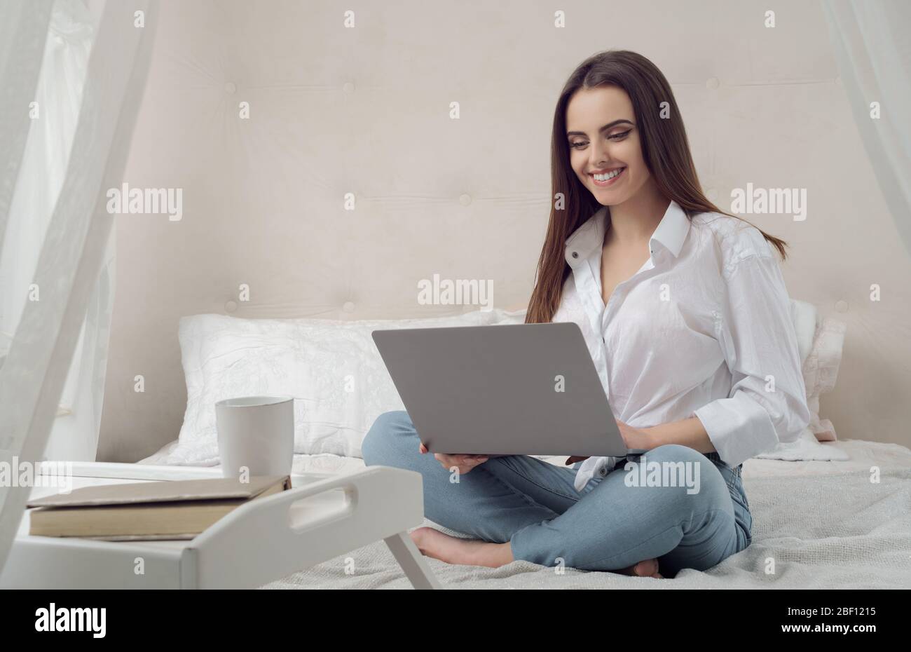 Happy Girl mit Notebook während des Sitzens auf bed.Young casual Mädchen arbeitet auf Laptop von ihrem Bett während der Quarantäne und lächelnd Stockfoto