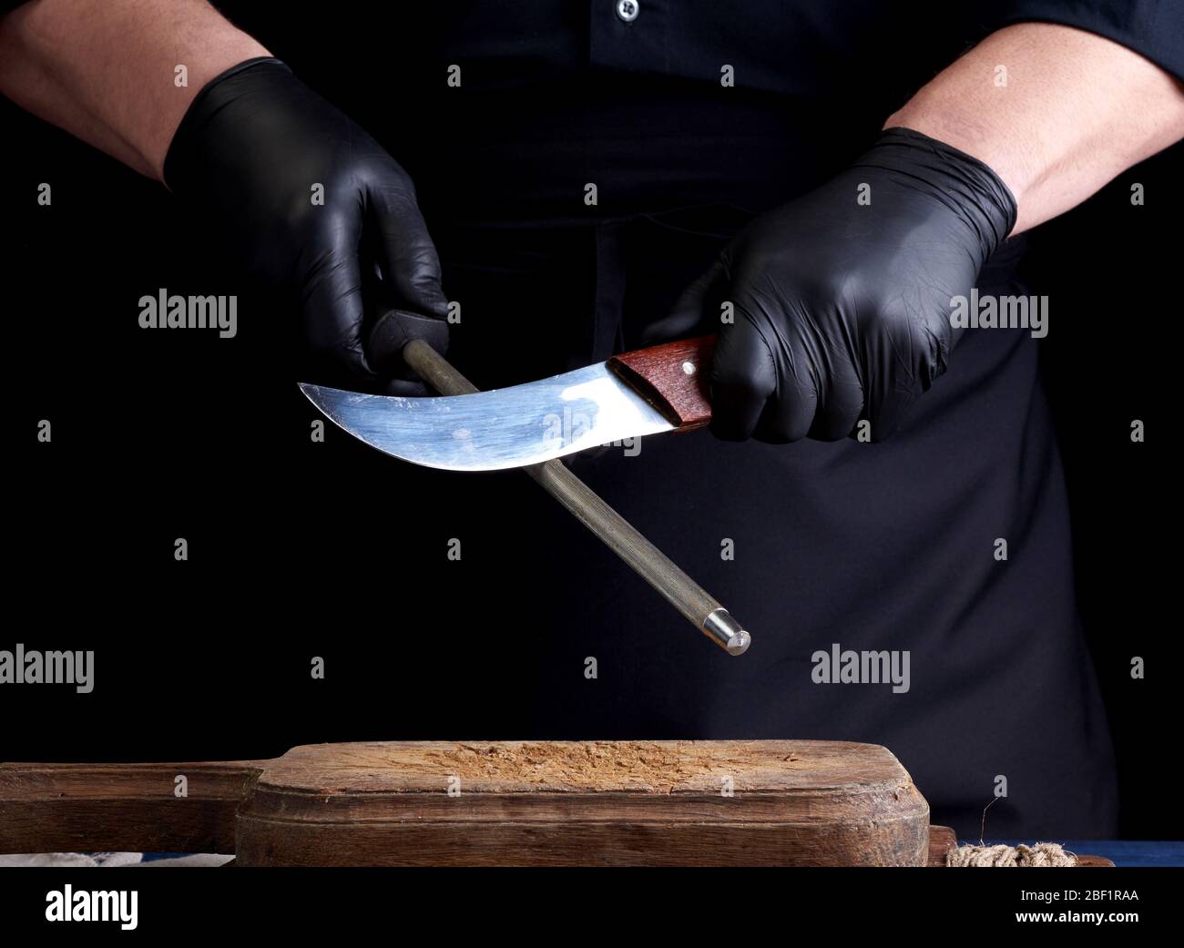 Koch in einem schwarzen Hemd und schwarzen Latex Handschuhe Messer auf einem eisernen Spitzer mit einem Griff über den Tisch, Low Key schärfen Stockfoto