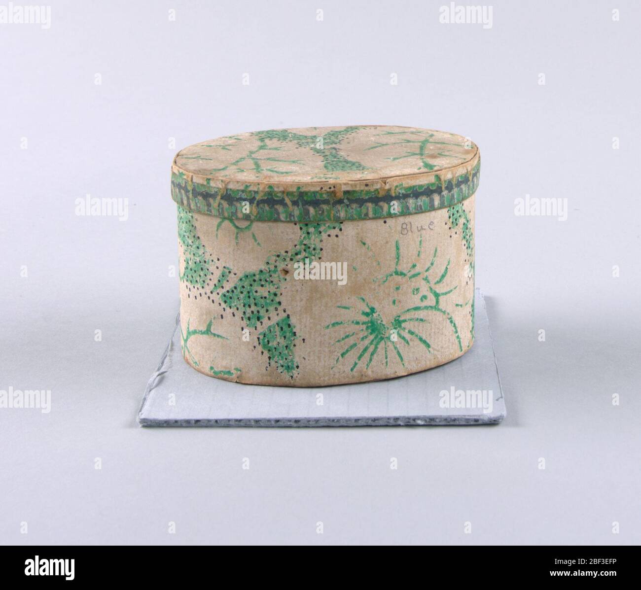 Bandbox. Sehr kleine ovale Bandbox. Papier ist wahrscheinlich Tapete: Kalkweiß-weiße Blüten und Blätter in hellgrün gepflückt. Kleine schwarze Punkte sind über einen Teil des Designs gestreut. Der Deckel ist mit einem geriffelten Design eingefasst, mit dunkelgrau schattiert. Stockfoto