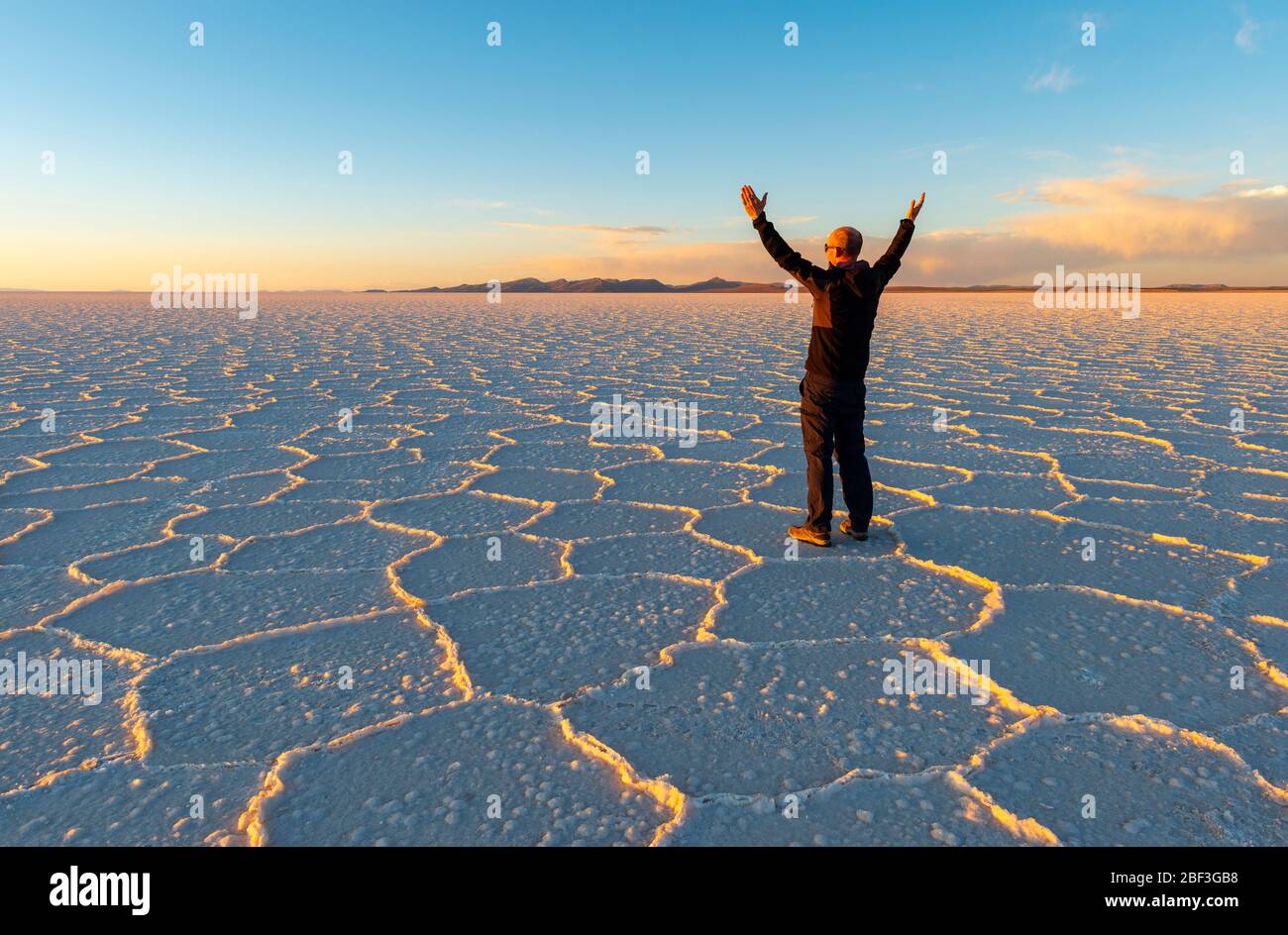 Sonnenuntergang der Uyuni Salt Flat Desert mit Hexagon Salzformationen und einem kaukasischen männlichen Touristen mit gestreckten Händen in der Luft, Bolivien. Stockfoto