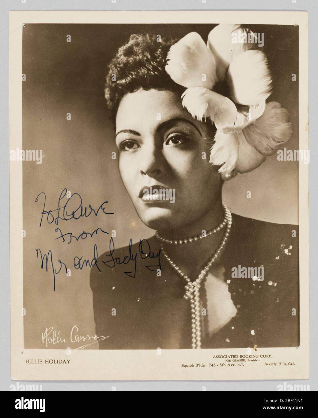 Foto von Billie Holiday. Ein Silbergelatinedruck, der ein Schwarz-Weiß-Bild von Billie Holiday zeigt. Der Feiertag steht hinter einem Mikrofonständer. Sie trägt eine weiße Bluse, einen Gürtel und einen Blumenrock. Im Hintergrund hinter ihr, auf der linken Seite des Bildes, ist ein Band abgebildet. Stockfoto