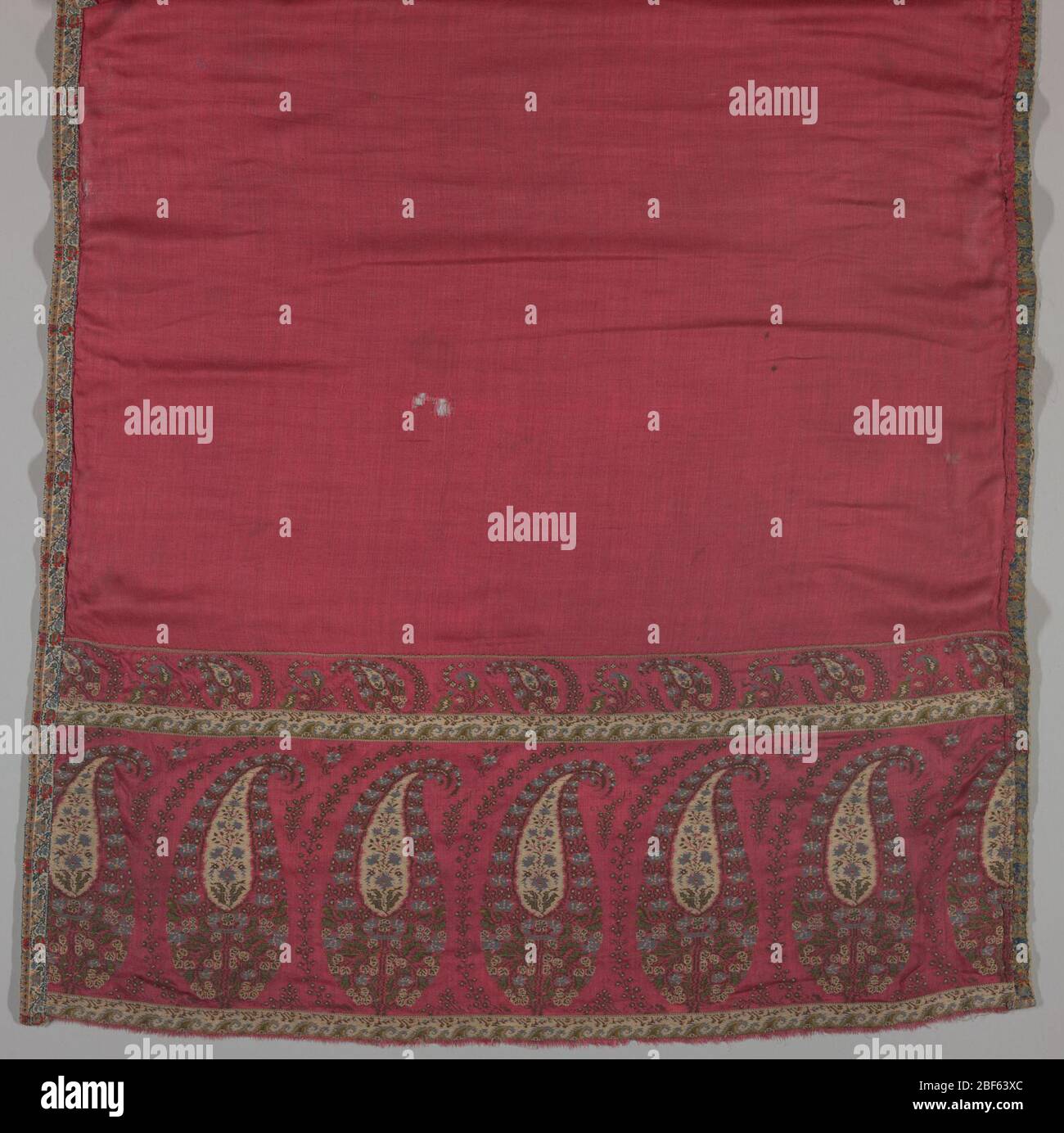 Schal. Paisley-Dessin auf rotem Tuch. Sechs große Kegel Motive an beiden Enden des Schalenschals. Einfarbige rote Seide wurde an den Paisley-Bordüren genäht. Ein Brokatband ist rund um den Schal genäht. Hellblau, grün, dunkelbraun und rot sind die vorherrschenden Farben. Stockfoto
