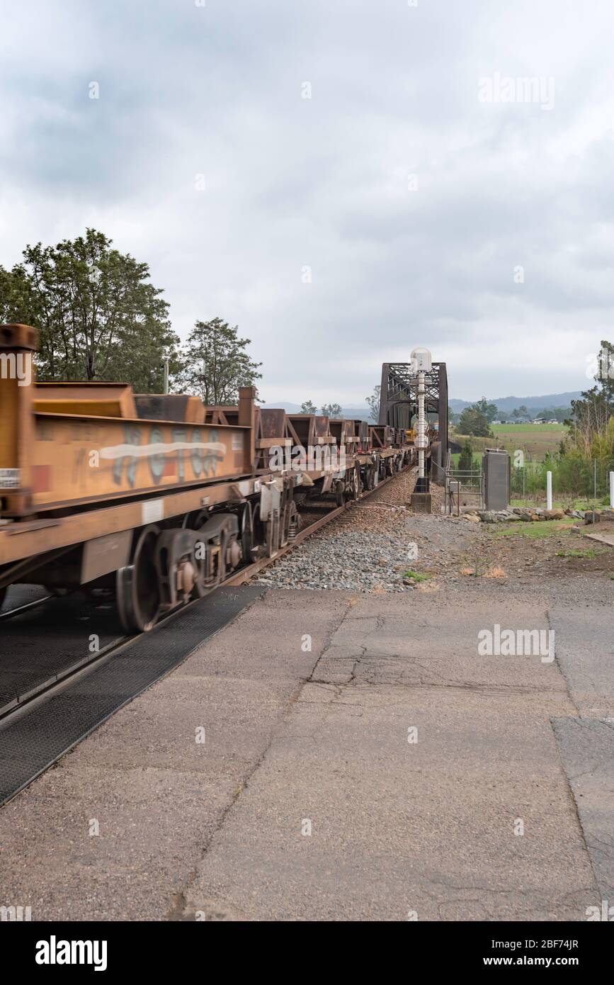 Eine Linie von Flachwagen, die von Diesellokomotiven an einem Bahnübergang durch die ländliche Stadt Paterson, New South Wales, Australien, abgeschleppt werden Stockfoto