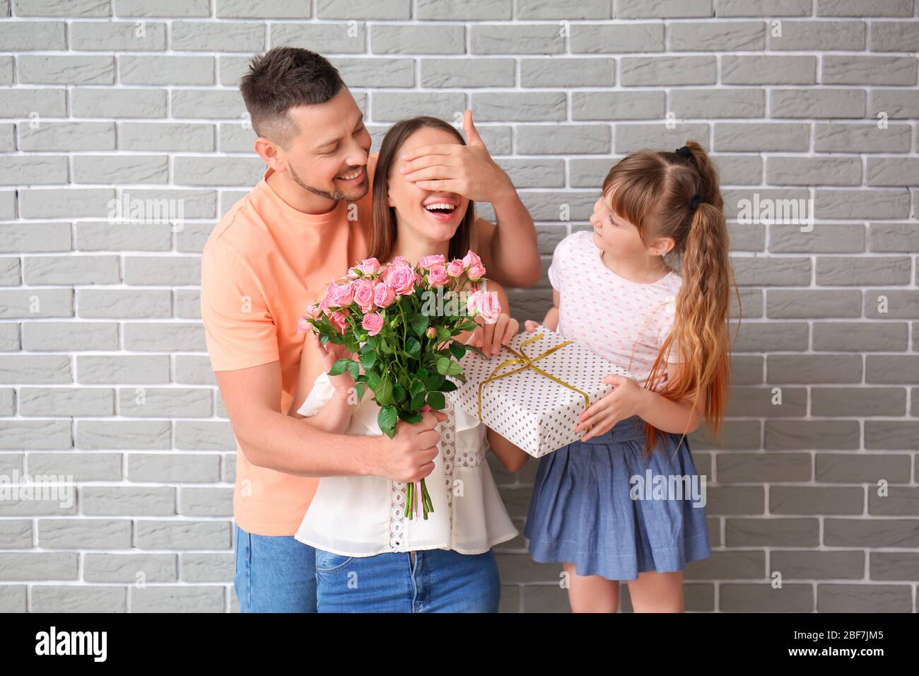 Ehemann und kleine Tochter grüßen Frau zum Muttertag auf grauem Backstein Hintergrund Stockfoto