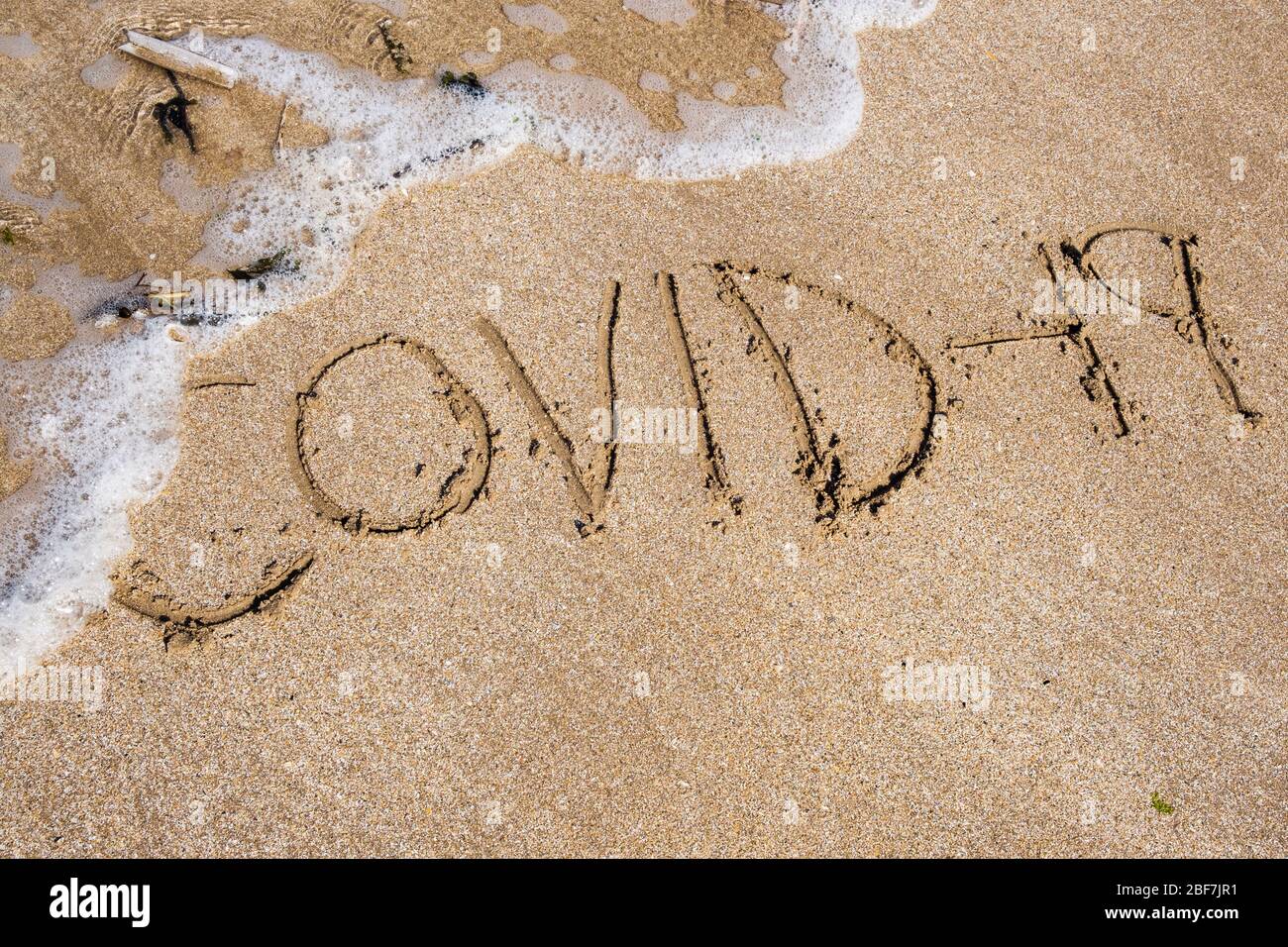 Covid-19 geschrieben in Sand, der von eingehender Flut weggespült wird. Zur Veranschaulichung des Konzepts der Coronavirus-Pandemie. Großbritannien Stockfoto