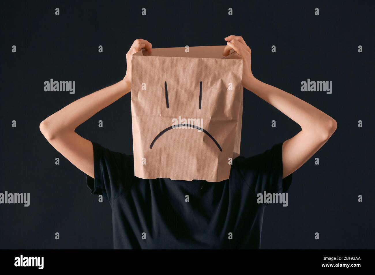 Frau trägt Papiertüte mit gezeichnetes trauriges Gesicht auf ihrem Kopf vor dunklem Hintergrund. Konzept der Depression Stockfoto