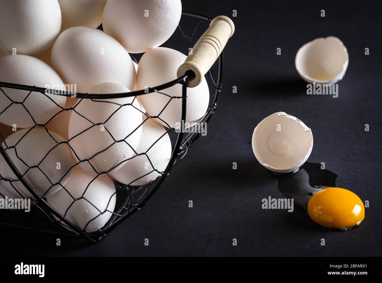 Hühnereier in einem Metallkorb auf einem schwarzen Holztisch. Rissige Ei direkt auf dem Tisch mit dem gelben Eigelb und gebrochene Eierschale. Konzeptbild für den Stockfoto