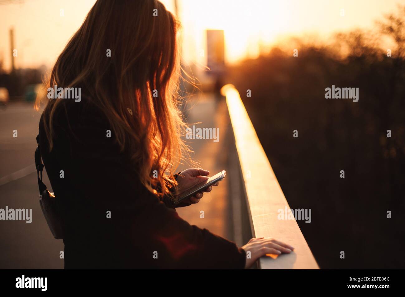 Junge Frau mit Smartphone während der Geländer auf der Brücke in der Stadt bei Sonnenuntergang Stockfoto