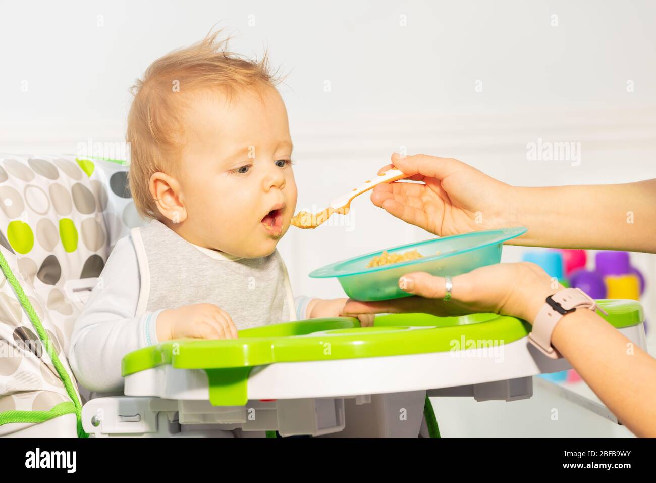 Kleines Kleinkind Baby Junge sitzen in Hochstuhl eifrig, Essen aus Löffel von Mutter vorgeschlagen essen Stockfoto