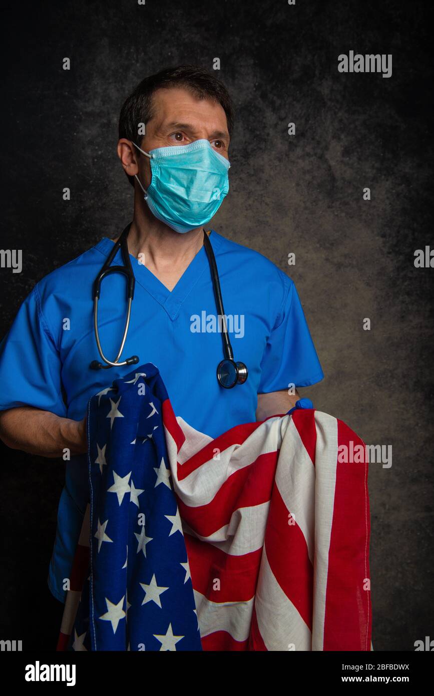 Arzt sucht nach Hoffnung/Heilung, trägt blaue Krankenhauspeelings mit Gesichtsmaske und Stethoskop, hält die amerikanische Flagge wie ein Baby in den Armen. Stockfoto