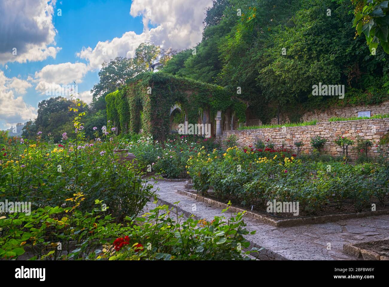 Schöner romantischer Garten mit bunten Blumen, Bögen mit grünen Pflanzen, Steinmauern und Gassen überwuchert. Blauer Himmel und flauschige Wolken Hintergrund. Stockfoto