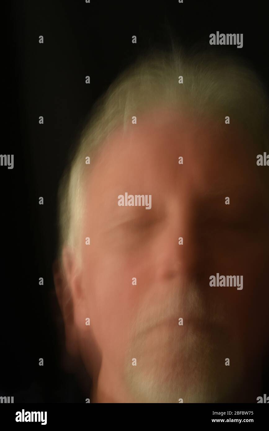 Das verschwommene Gesicht eines älteren Mannes mit weißem Haar und einem Ziegenbart mit in einem meditativen Zustand geschlossenen Augen vor schwarzem Hintergrund. Stockfoto