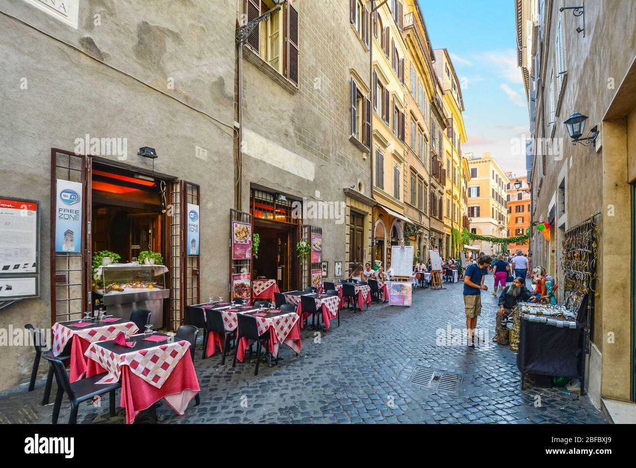 Buntes italienisches Restaurant mit Tischen in einem Straßencafé in einer Gasse in Rom, Italien, mit einem Straßenverkäufer, der Souvenirs an einen Kunden verkauft. Stockfoto