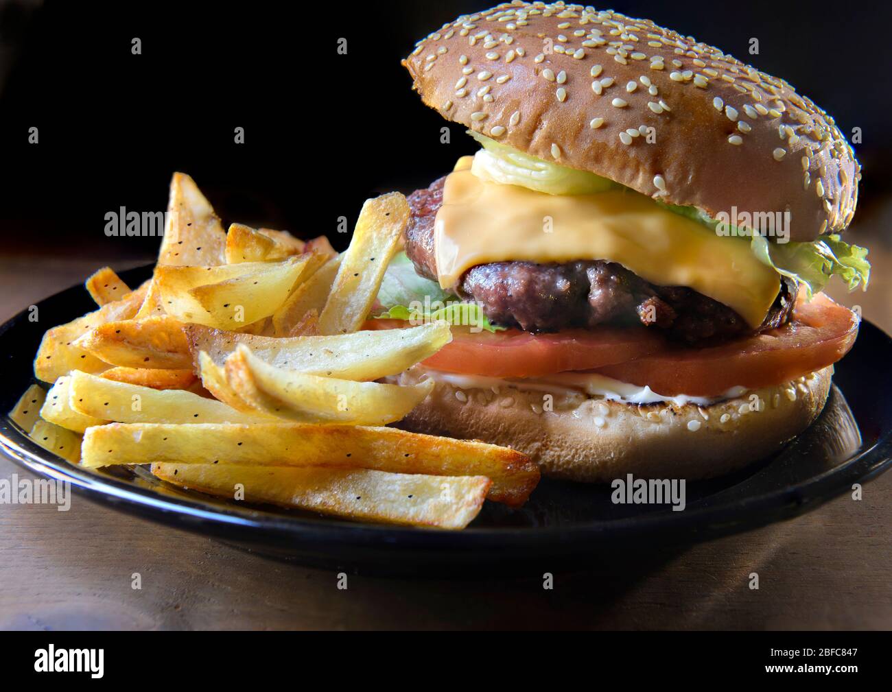 Ein hausgemachter Hamburger, mit pommes frites an der Seite, auf einem rustikalen Holztisch und mit Randlicht auf dem Hamburger, leckere Speisen Stockfoto