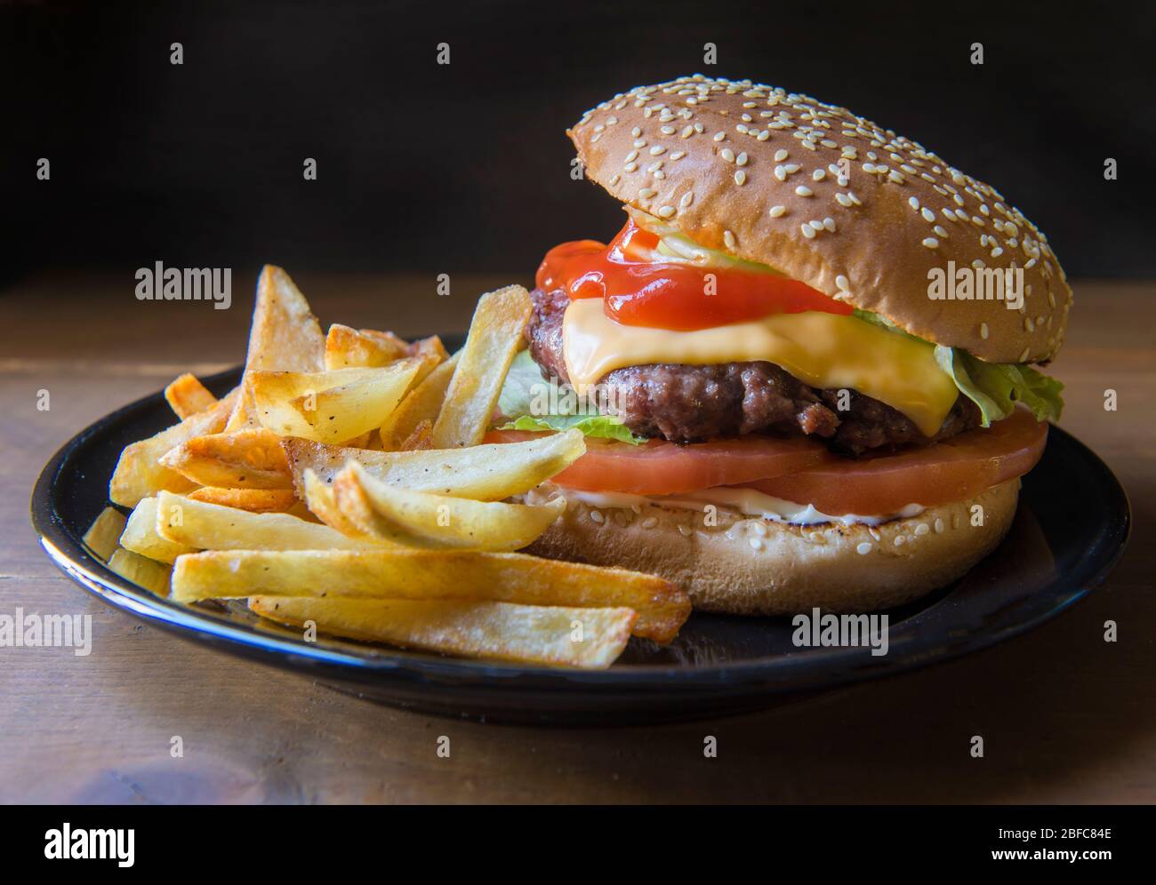 Hausgemachter Hamburger mit Ketchup und pommes frites auf der Seite auf einem schwarzen Teller auf einem rustikalen Holztisch, mit schwarzem Hintergrund, leckere Speisen. Stockfoto