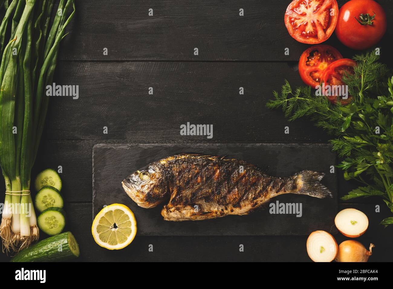 Gekochter dorado-Fisch auf schwarzem Stein. Schwarzer Holzhintergrund mit Tomaten, frischen Zwiebeln und anderem Gemüse Stockfoto