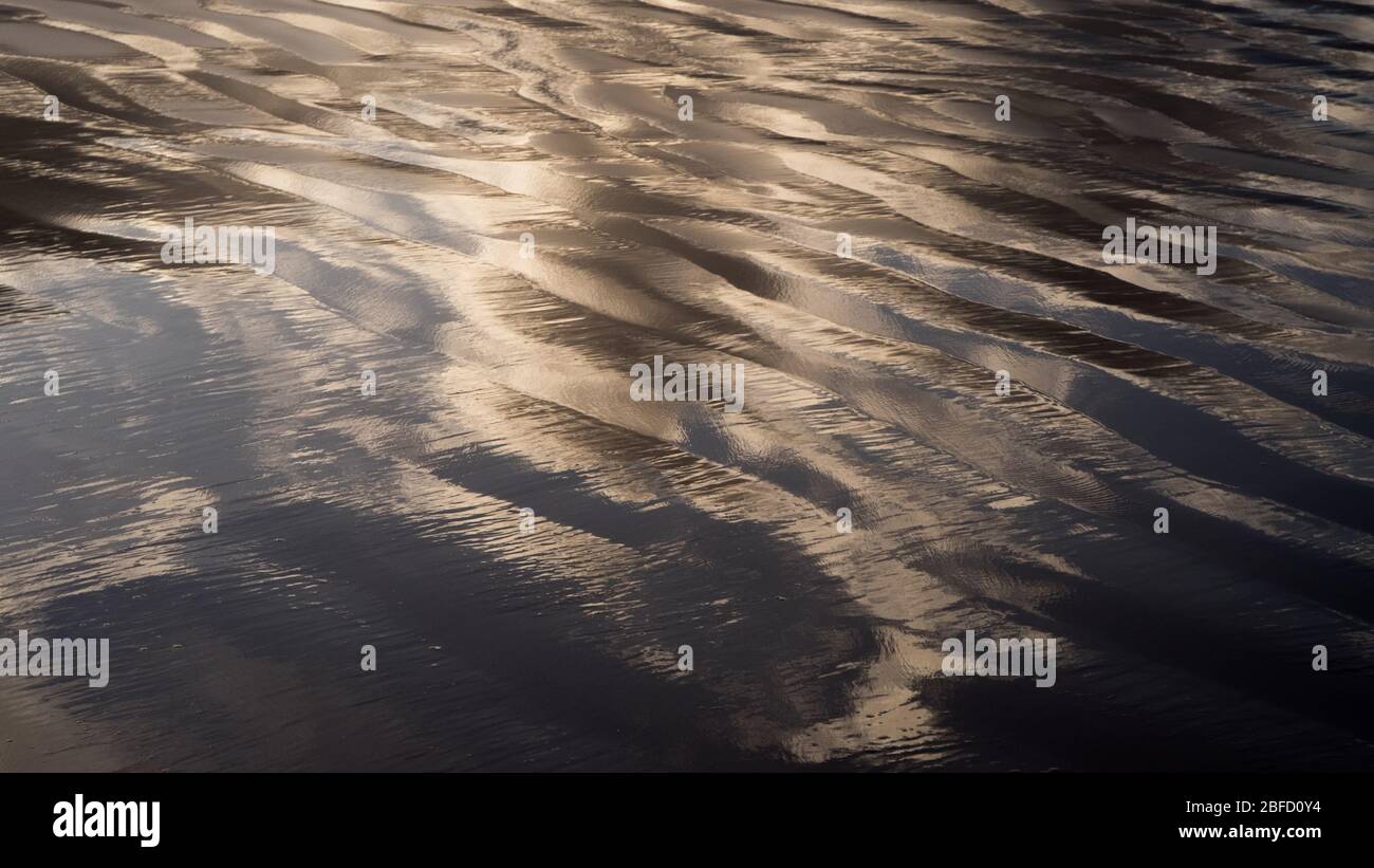 Eine abstrakte Nahansicht der nassen Sandfläche am Strand mit kleinen Pfützen und Spiegelung von dunkelgrauen Wolken Stockfoto