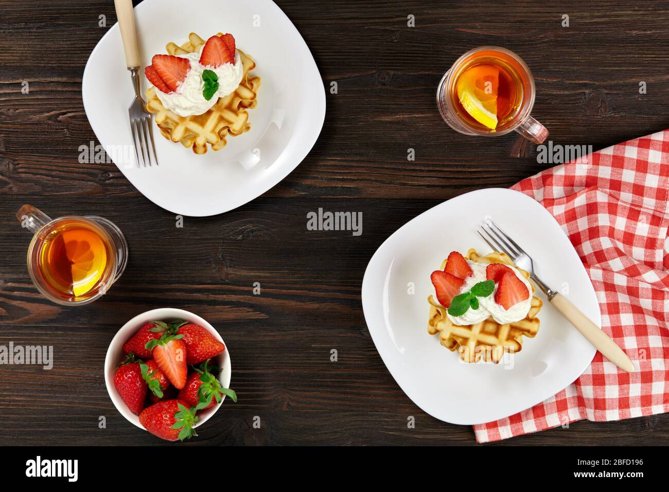 Wunderschönes Frühstück. Belgische Wiener Waffeln mit Sahne und Erdbeeren, Tee und karierter Serviette auf dunklem rustikalem Holzhintergrund. Stockfoto