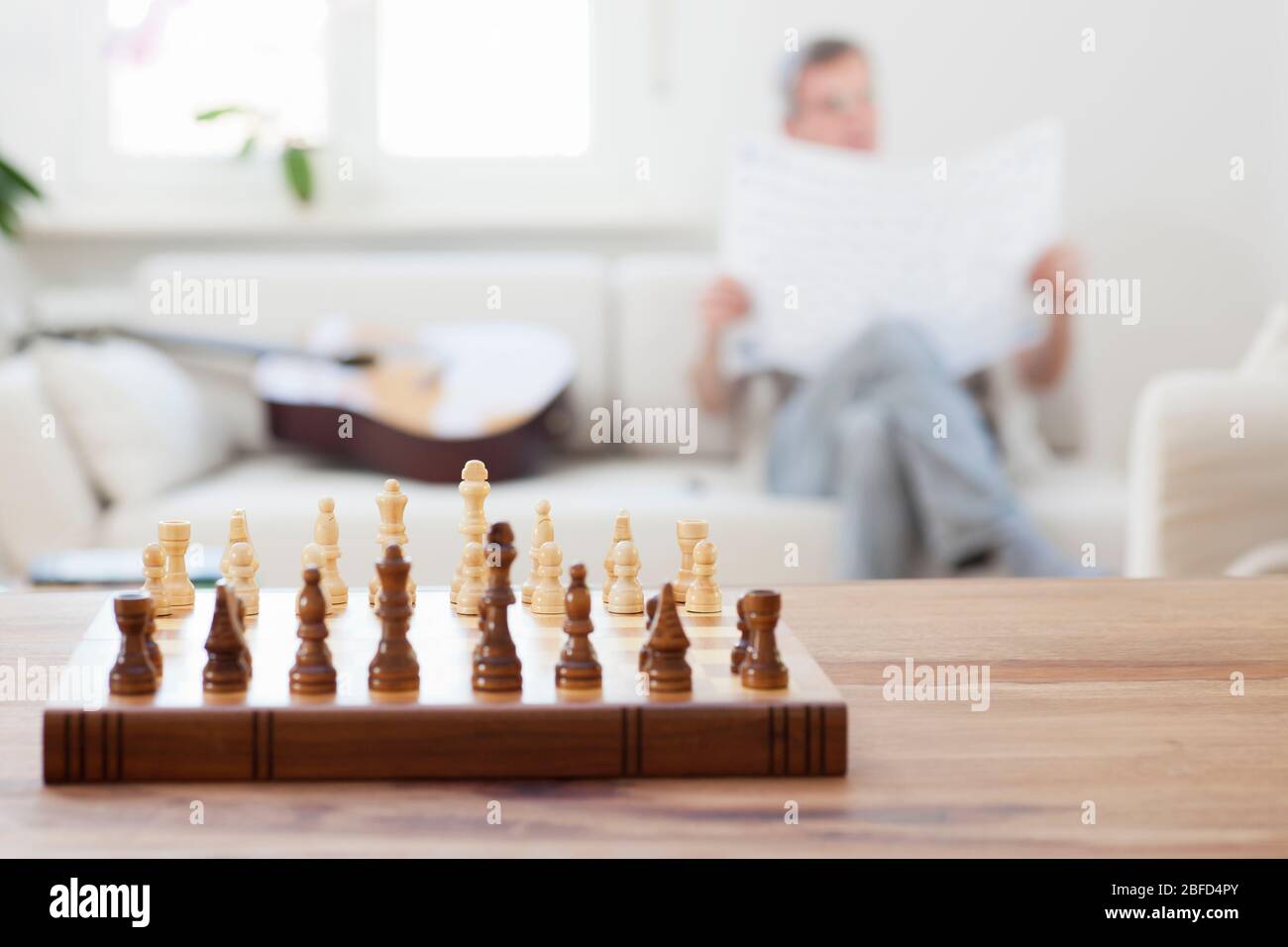 Senior liest Zeitung auf einer Couch in einem hellen Wohnzimmer mit Schachbrett im Vordergrund - Fokus auf das Schachbrett Stockfoto