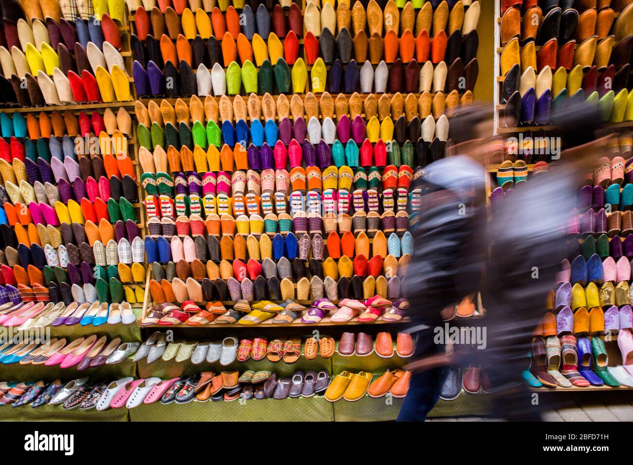Fußgänger schlendern an einem Straßenstand vorbei, an dem bunte traditionelle Lederpantoffeln in der Medina (Altstadt) von Fez, Marokko, verkauft werden. Stockfoto