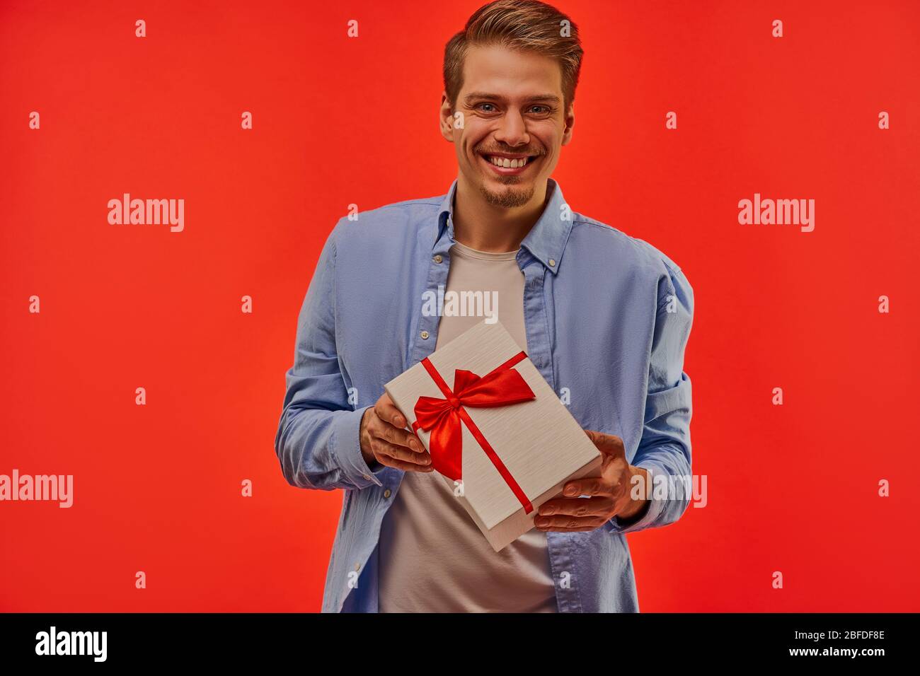 Ein Kerl lächelt weit mit weißen Kasten mit einer roten Schleife und Bändern in den Händen. Stockfoto